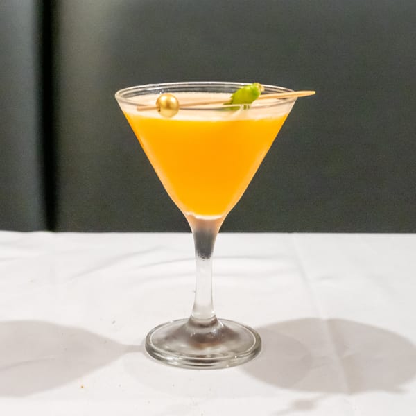 Bright orange cocktail in a stemmed glass, garnished with a green leaf and gold cocktail pick, on a white table.