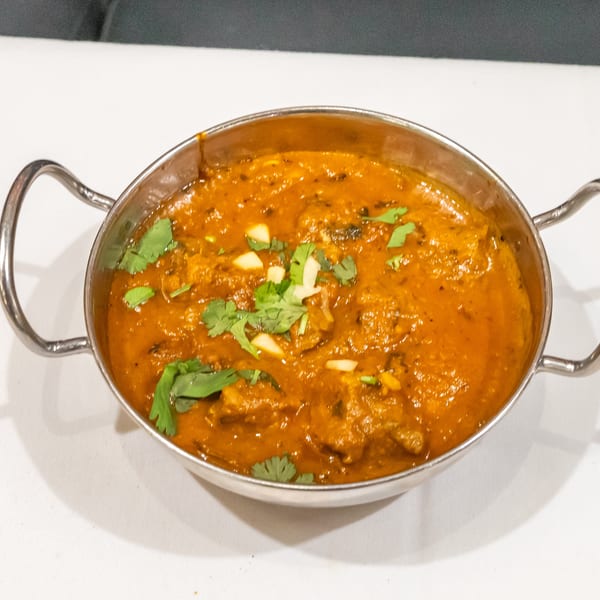 A metal serving bowl holds rich orange curry garnished with fresh cilantro and sliced garlic on a white tablecloth.