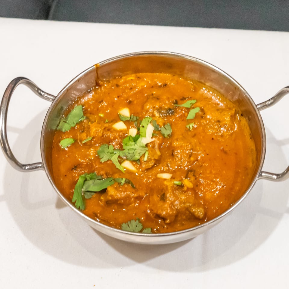 A metal serving bowl holds rich orange curry garnished with fresh cilantro and sliced garlic on a white tablecloth.