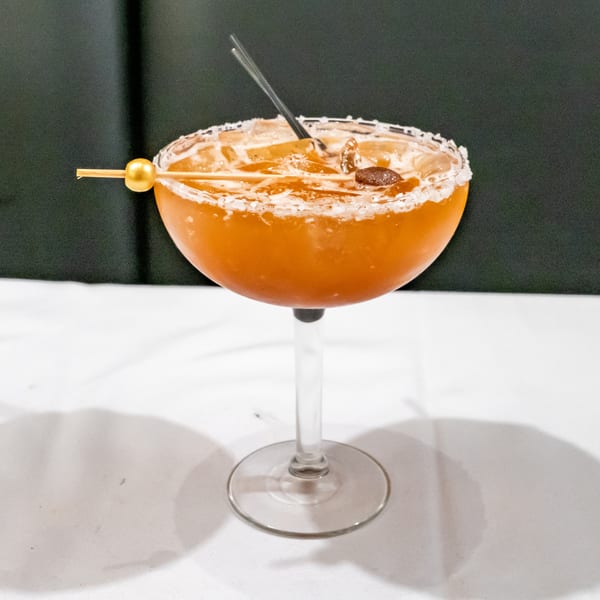 Amber cocktail in a coupe glass with a crushed-ice rim, cocktail pick garnish, and dark berry on a white table.