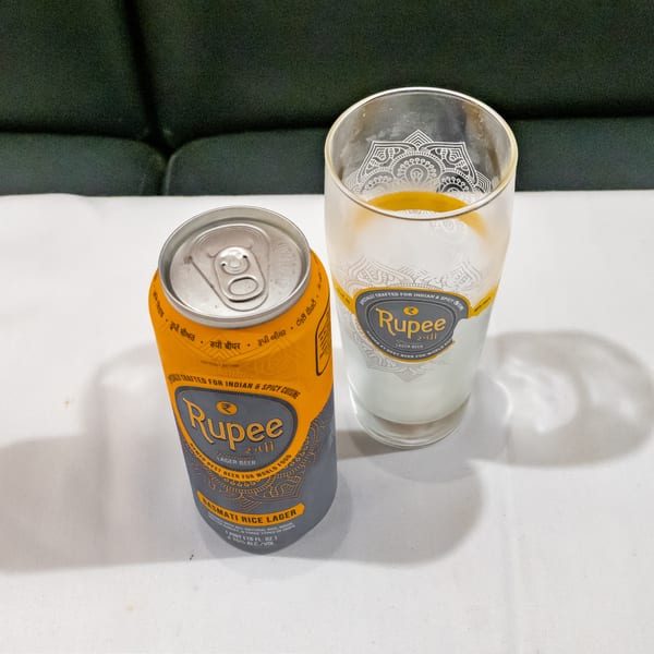 Rupee Premium Lager beer can and partially filled decorative beer glass on a white table beside dark upholstered seating.