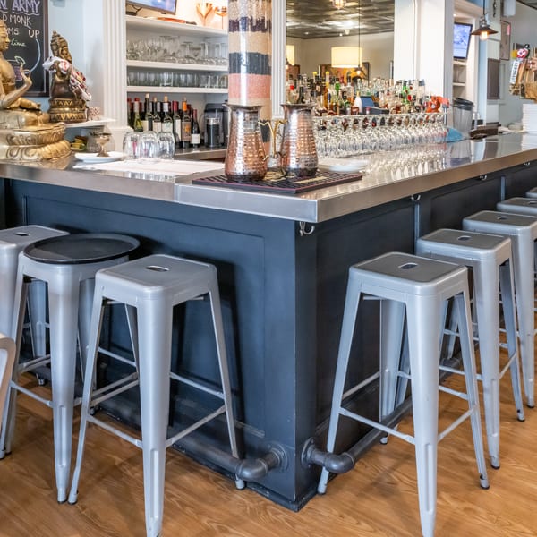 Restaurant bar with copper pitchers, rows of glassware, liquor bottles, Buddha statues, and gray metal stools.