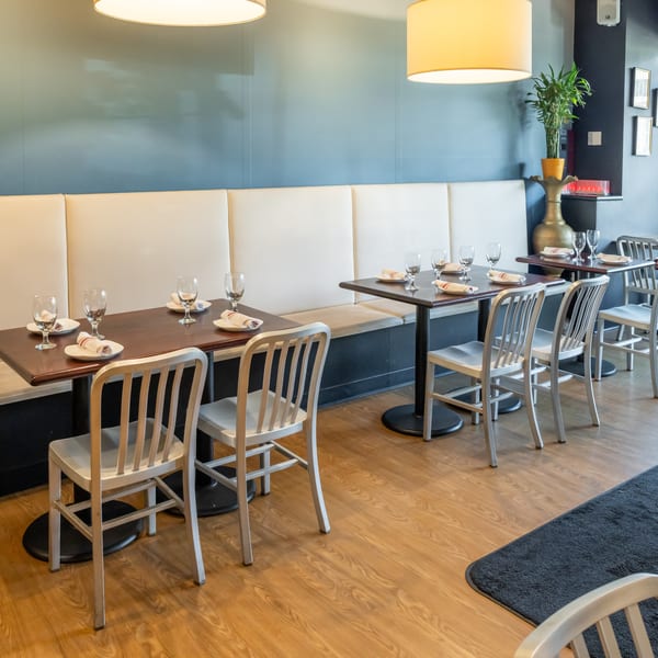 Bright restaurant dining room with cream banquettes, neatly set tables, metal chairs, pendant lights, and a potted plant.
