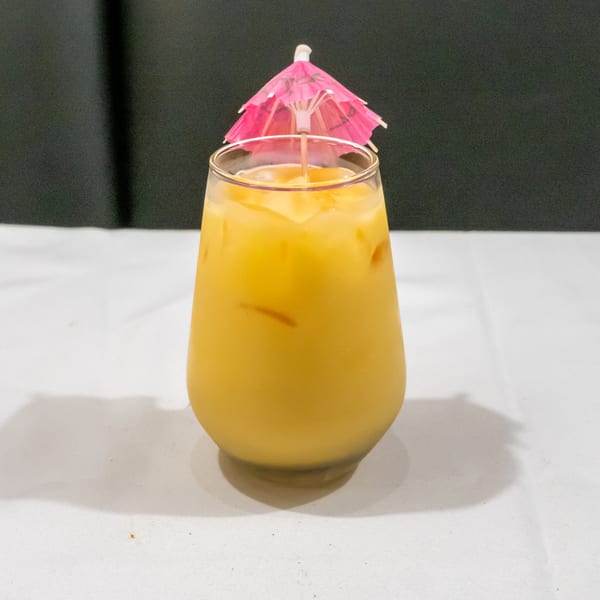Yellow iced cocktail in a rounded glass topped with a pink paper umbrella on a white table.