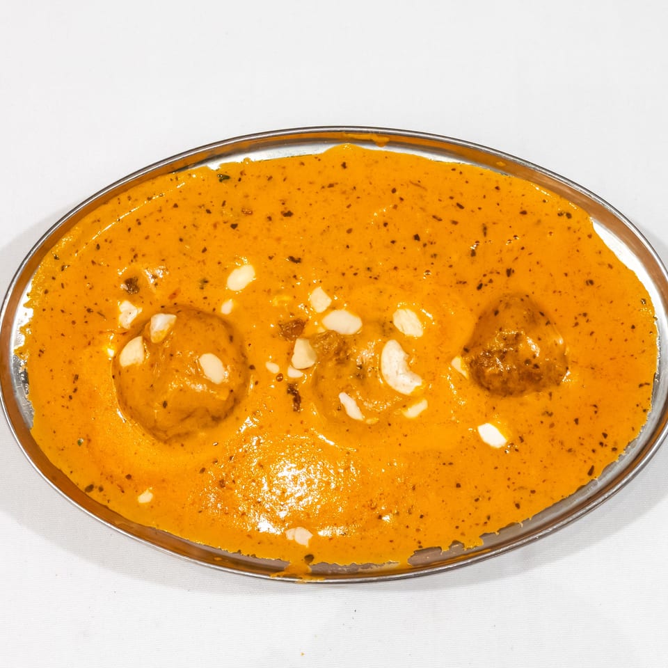 Creamy orange curry with meatballs, cashews, raisins, and herbs served in an oval metal dish.