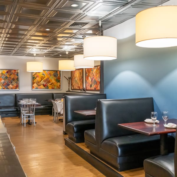 Elegant restaurant dining room with black leather booths, colorful artwork, pendant lamps, and set tables beneath a decorative ceiling.